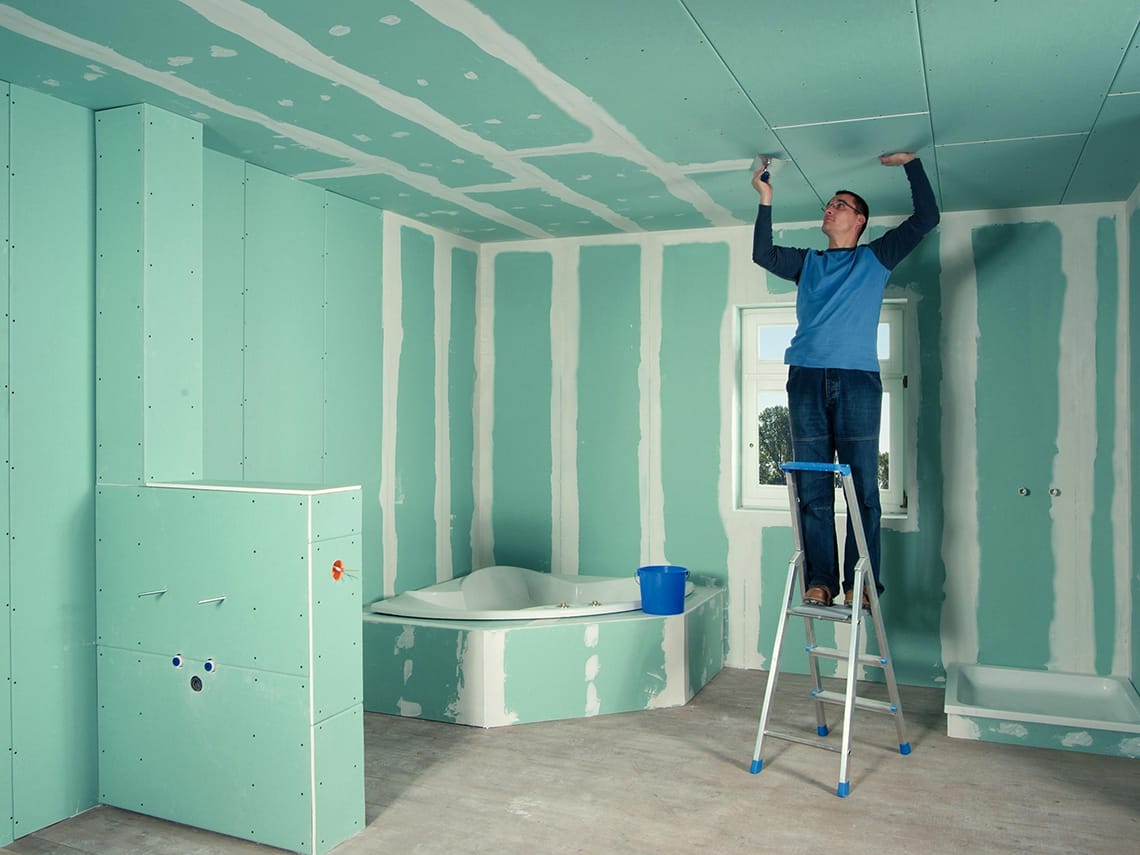 Ein Mann auf einer Leiter tätigt Trockenbauarbeiten an der Decke eines Badezimmers